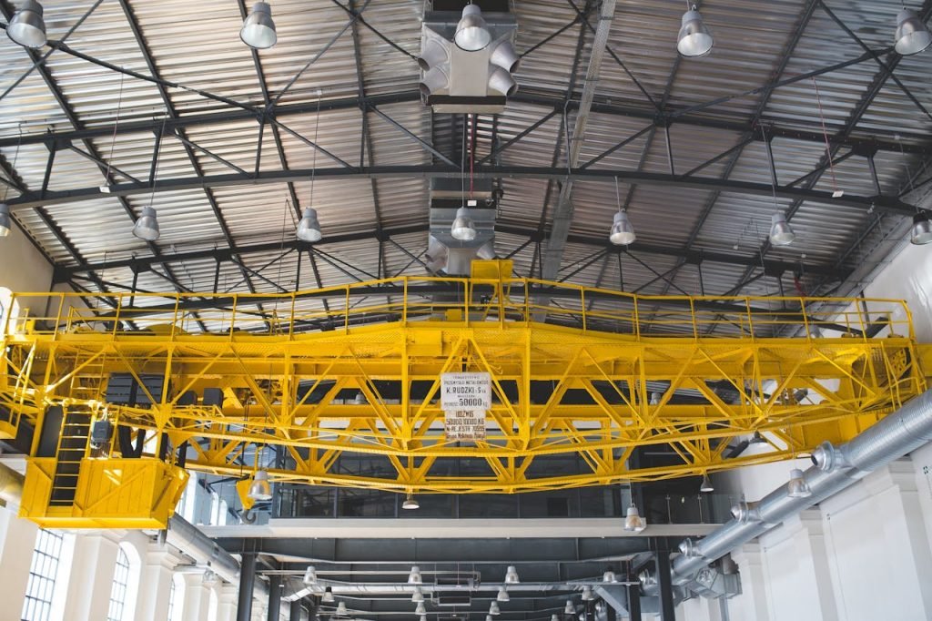 A yellow gantry crane inside an industrial factory with metal roof and lights.